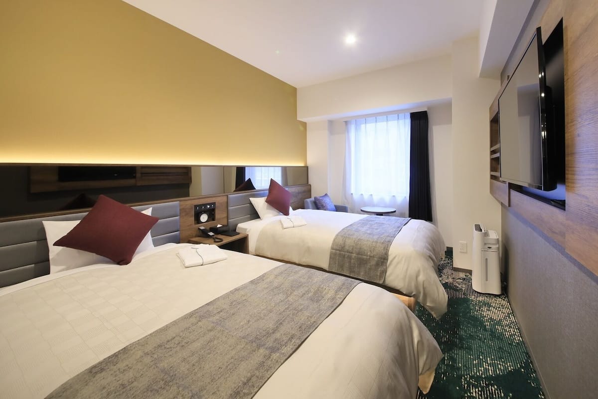 A hotel room with two beds, a flat-screen TV, a bedside table with a phone, a window with curtains, and a carpeted floor.