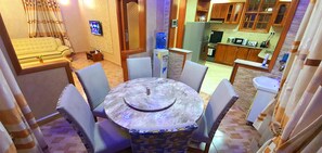 A dining area with a round table and six chairs, a sofa, a television, and a kitchen in the background. - Cape of Goodhope Vacation Home (Arusha)