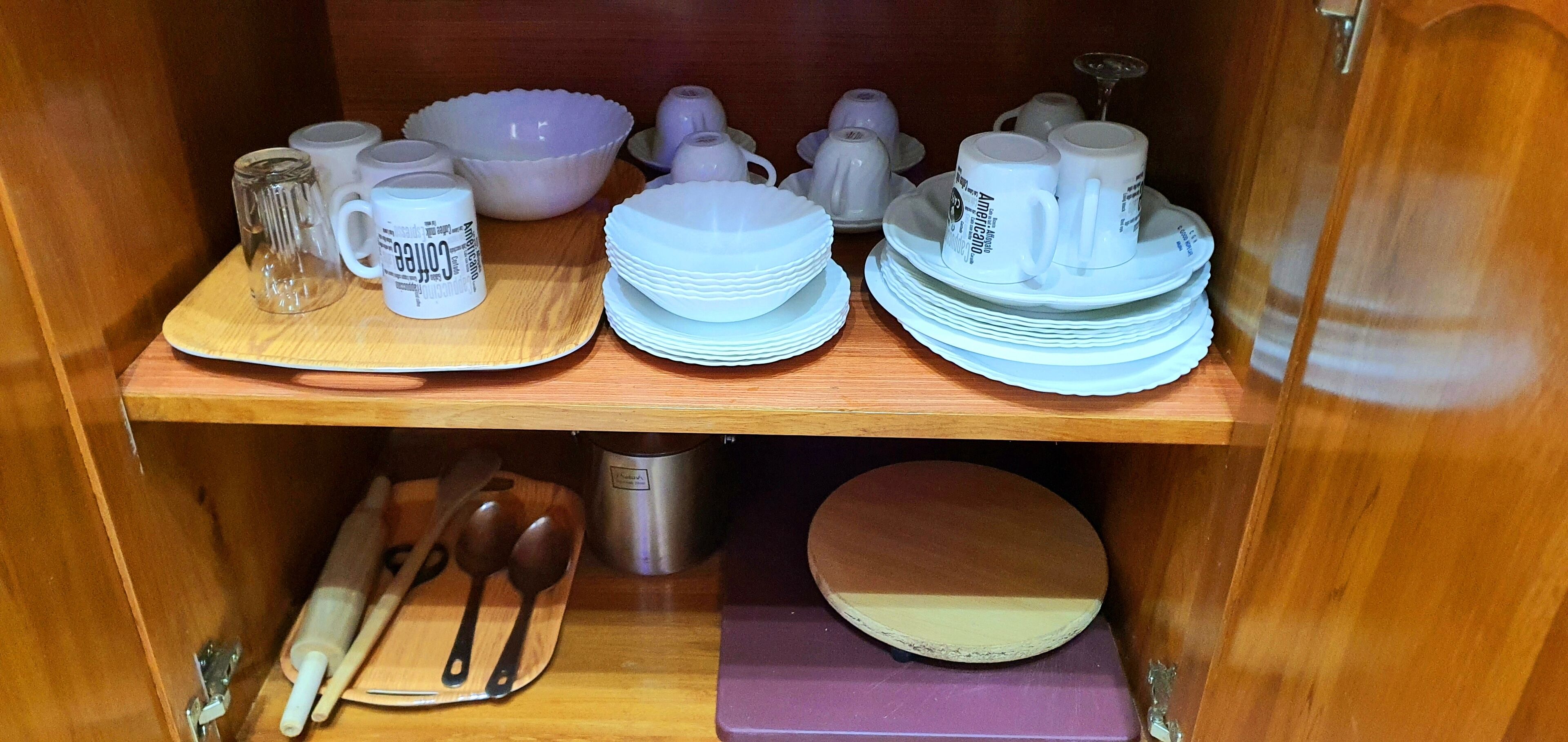 A wooden cabinet with shelves containing white ceramic mugs, plates, bowls, and a metal container.