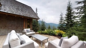A wooden cabin with a stone roof, surrounded by a patio with outdoor furniture and a view of mountains and trees. - Luxury chalet w/ hot tub & media (Whistler)