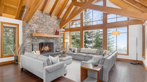 A spacious living room with a stone fireplace, large windows, and a high wooden ceiling. - Luxury chalet w/ hot tub & media (Whistler)