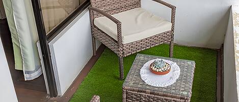 A small balcony with wicker furniture, a table with a decorative bowl, and artificial green grass.