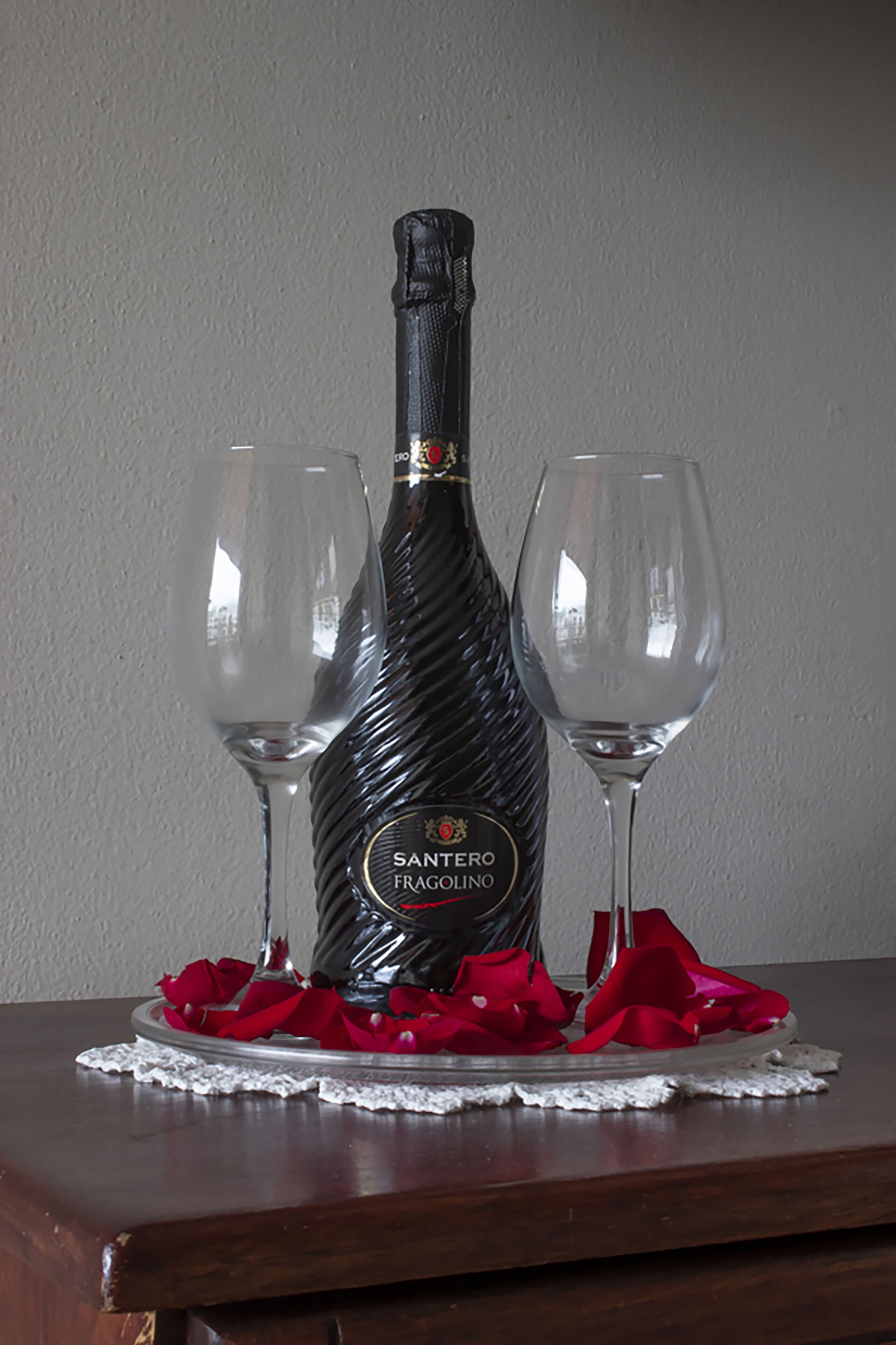 A bottle of Santérino Fragólino wine with two empty glasses on a wooden table, surrounded by red rose petals.