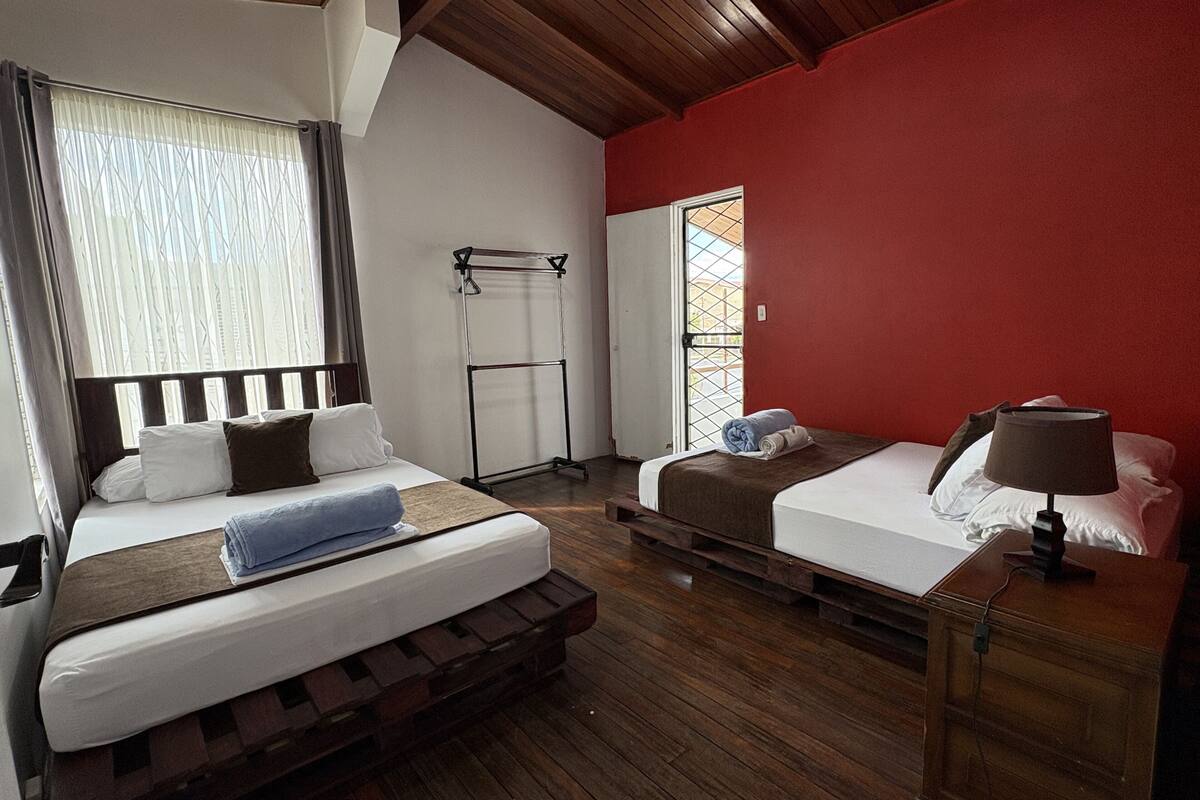 A room with two beds, a wooden floor, a red wall, and a window with curtains.