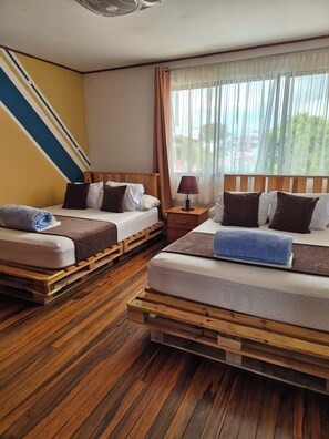 Ein Hotelzimmer mit zwei Betten, Holzboden, einem Fenster mit leichtem Vorhang und einem Deckenventilator.