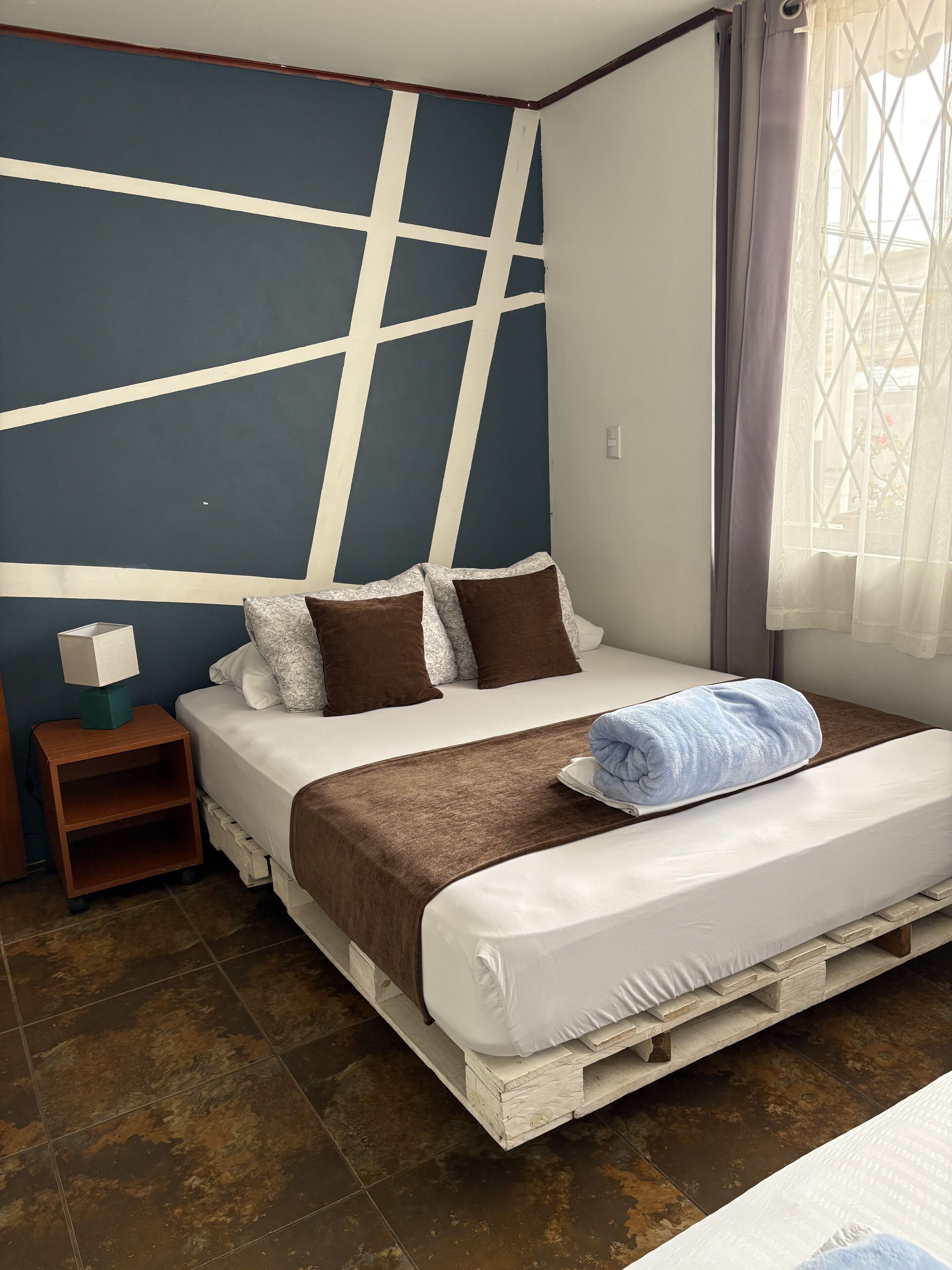 Un dormitorio con cama, almohadas, una mesita de noche y una ventana con cortinas.