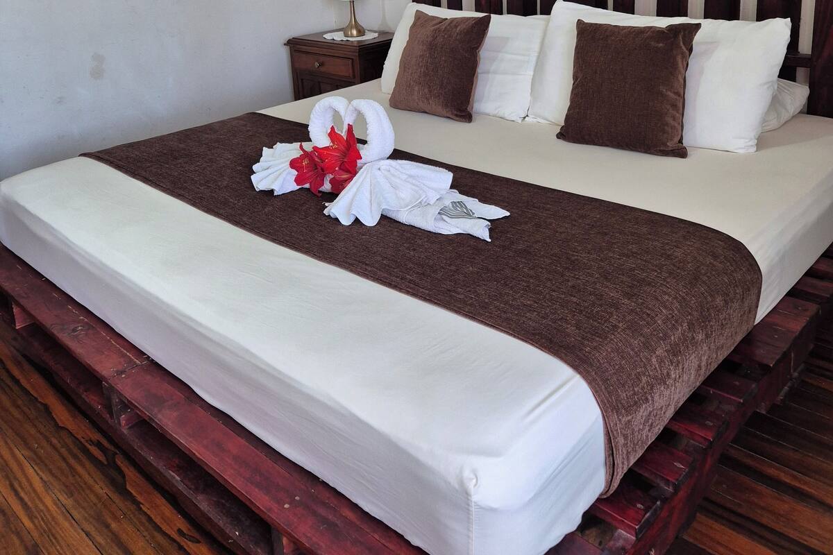 A hotel room with a wooden bed frame, white bedding, a brown throw blanket, and a decorative swan with red flowers.