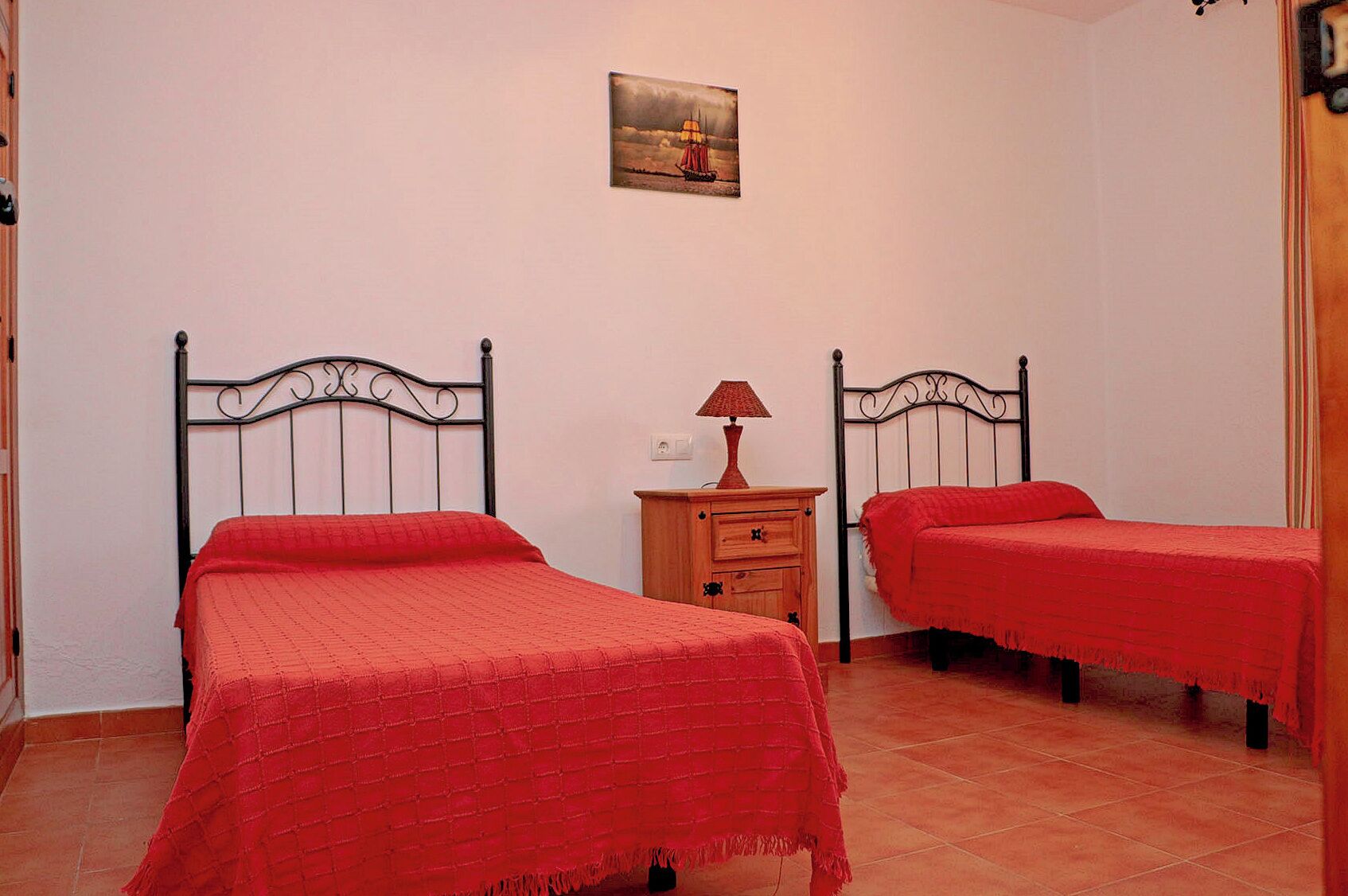 Een slaapkamer met twee bedden, elk met een rode sprei, een houten nachtkastje met een lamp en een lijst met een afbeelding aan de muur.