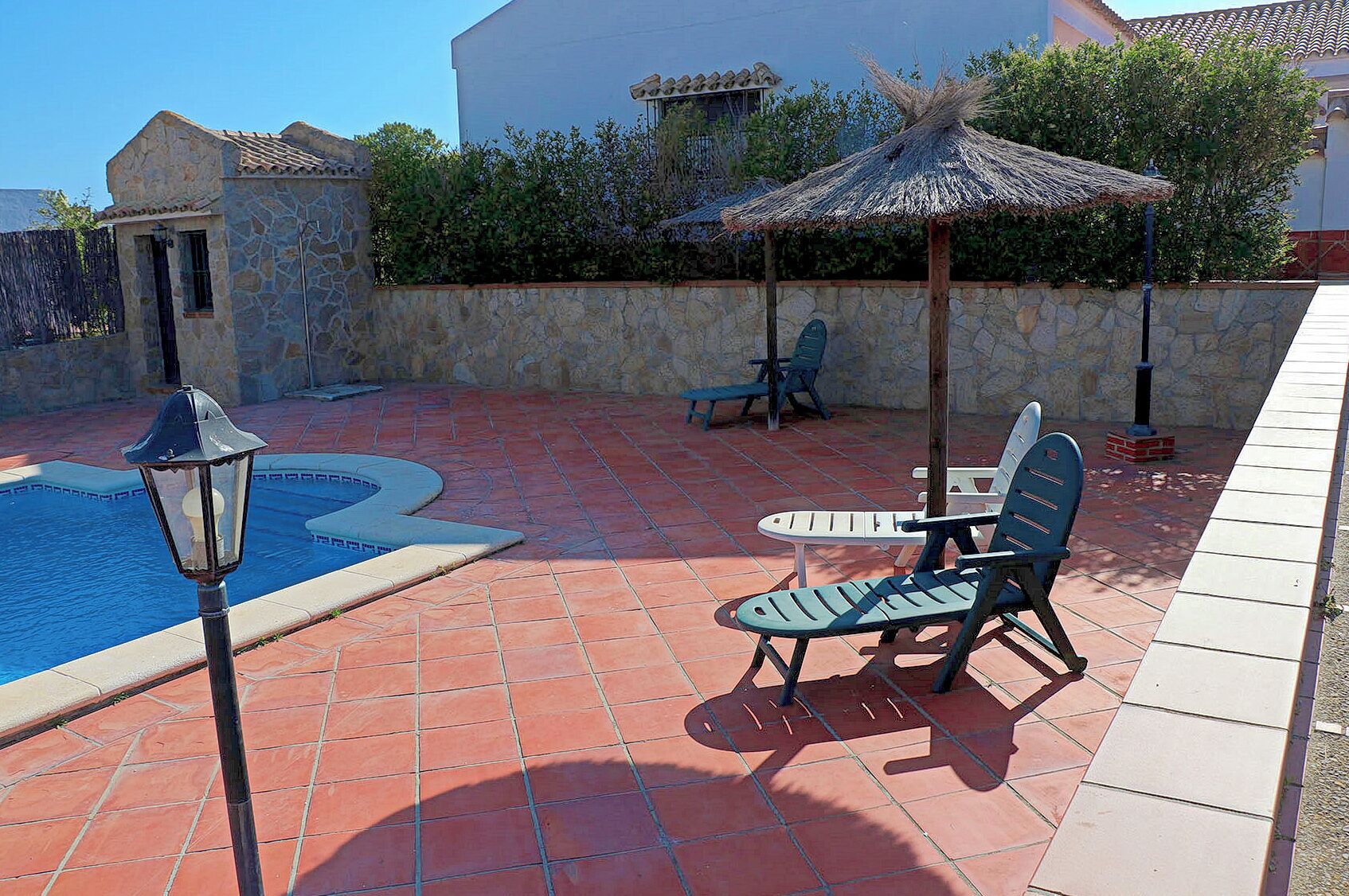 Una piscina con un muro de piedra, un área de descanso con sillas y una mesa, y una sombrilla de paja.