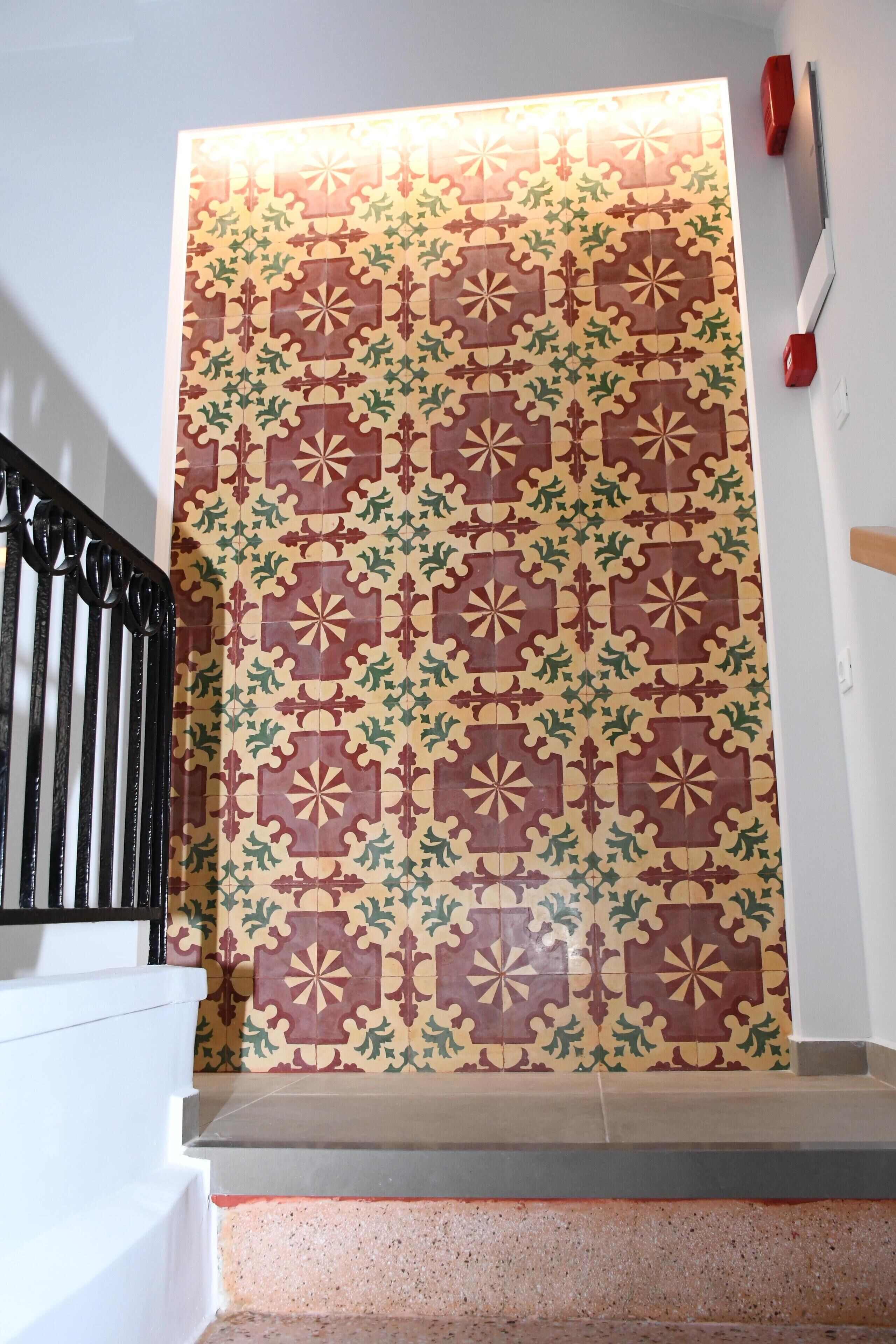 Una escalera con un muro de azulejos estampados, barandilla negra y una herramienta de mango rojo en la pared.
