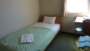 Una cama individual con una colcha verde y almohadas blancas, un escritorio pequeño con una silla, y una ventana con cortinas.