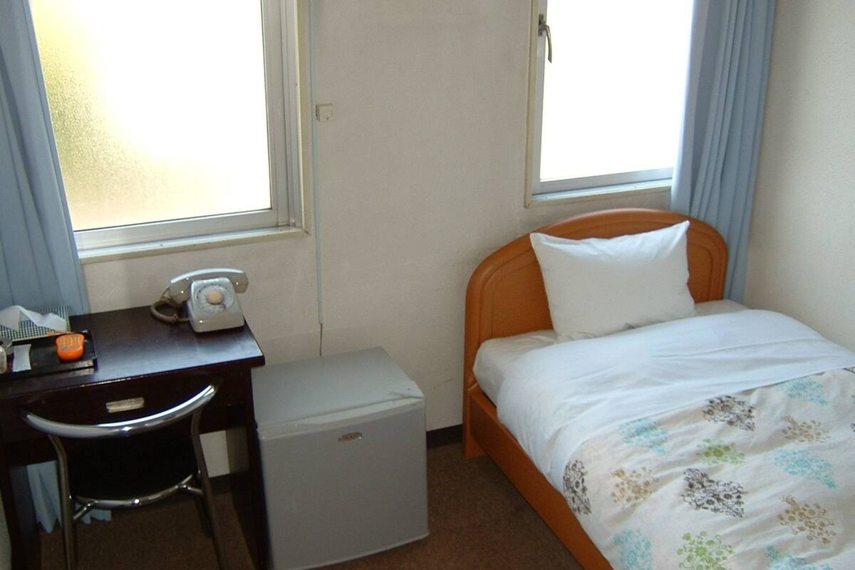 Una habitación de hotel pequeña y sencilla, con una cama, un escritorio con teléfono y un refrigerador compacto.