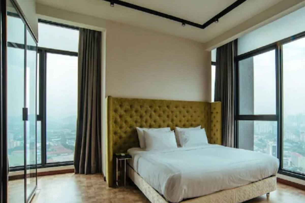 A modern bedroom with a large bed, a nightstand, and a view of the city through large windows.