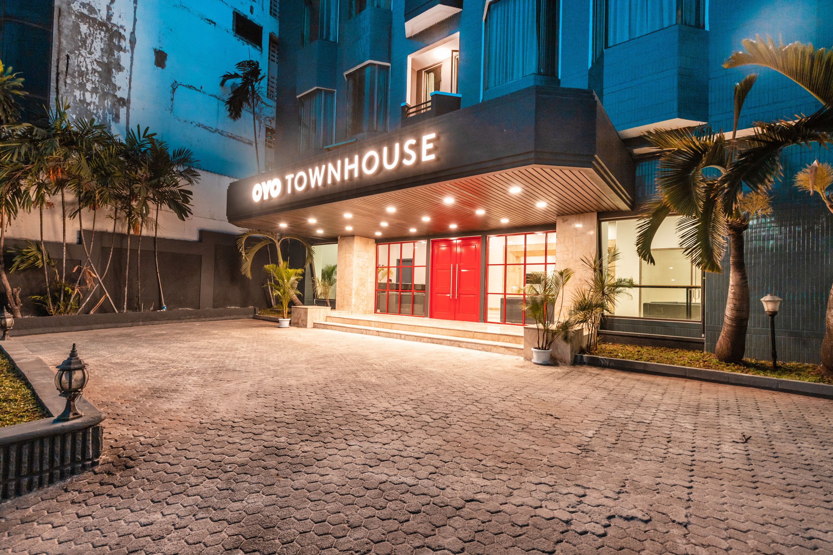 A hotel entrance with a red door, paved walkway, and palm trees.