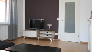 A modern living room with a flat-screen TV on a wooden TV stand, a lamp, and a door with frosted glass panels. - Apartment Anita Fischbach near Dahn (Fischbach bei Dahn)