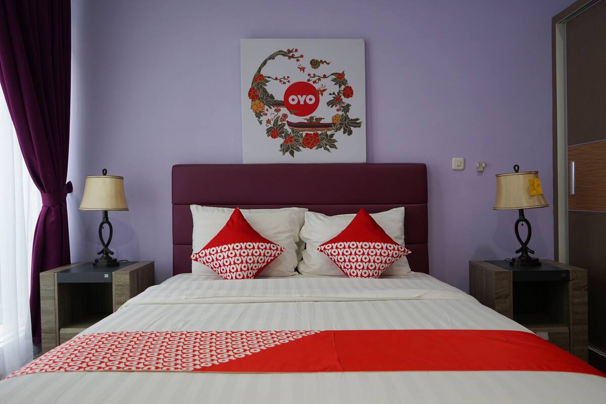 A neatly made bed with white and red bedding, two red pillows with a pattern, a wall-mounted artwork with the word "OYO", and two bedside lamps on wooden nightstands.