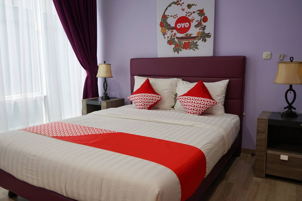 A hotel room with a bed, two bedside tables, a window with curtains, and a wall decoration.