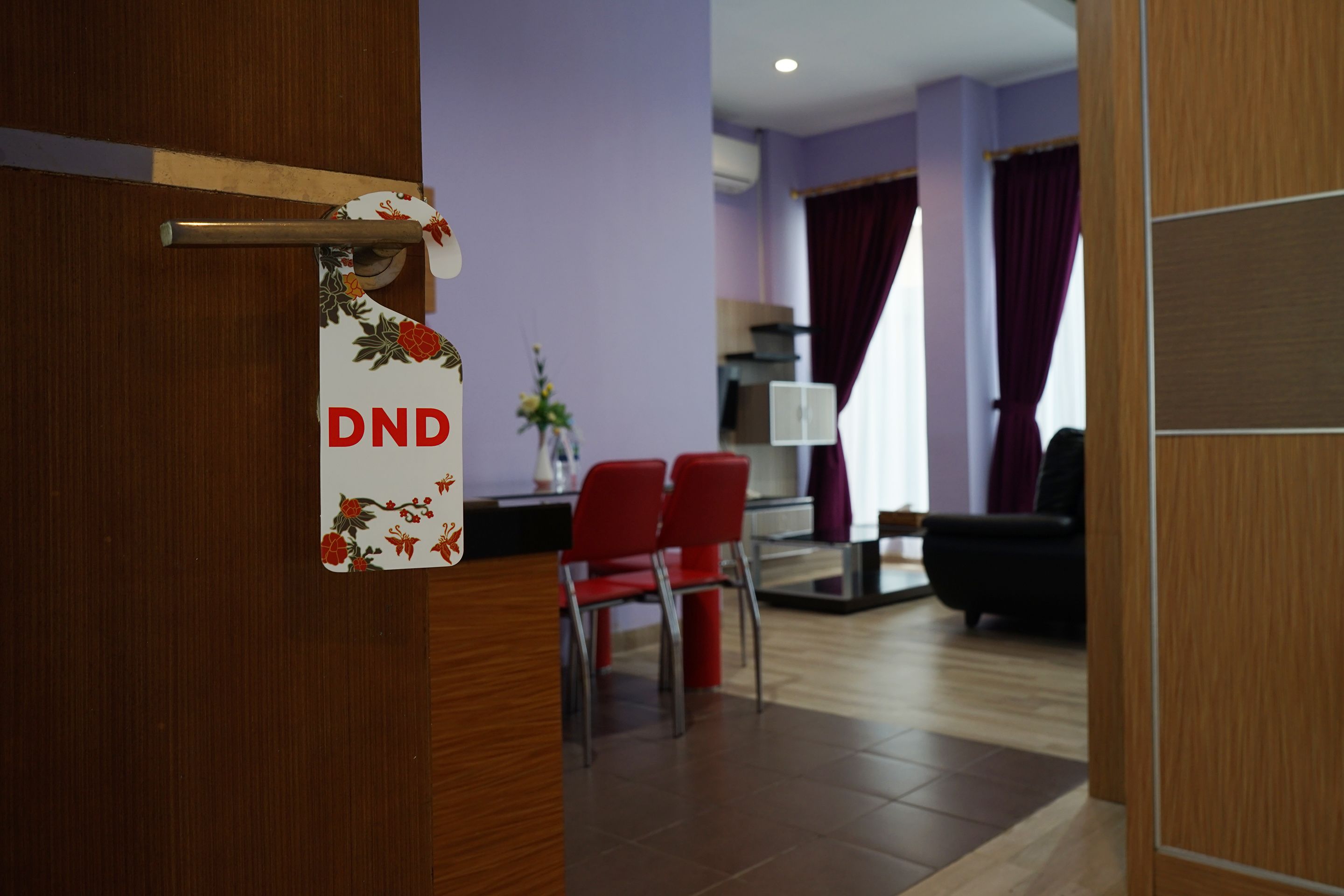 A hotel room with a wooden door, a red chair, a small table with a vase, and a view of the living area.