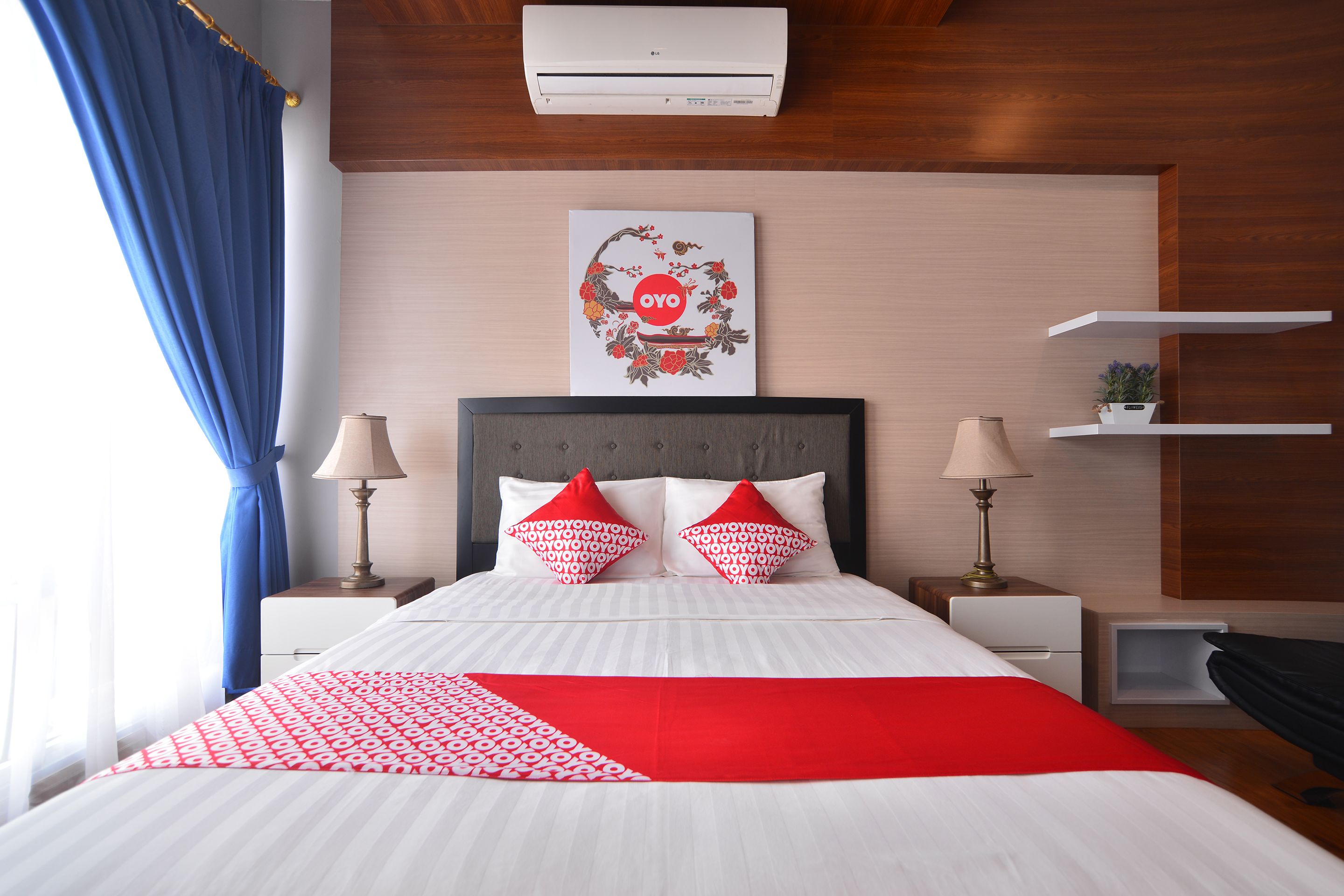 A neatly arranged hotel room with a bed, two bedside tables, a wall-mounted air conditioner, a decorative wall piece, and a blue curtain.