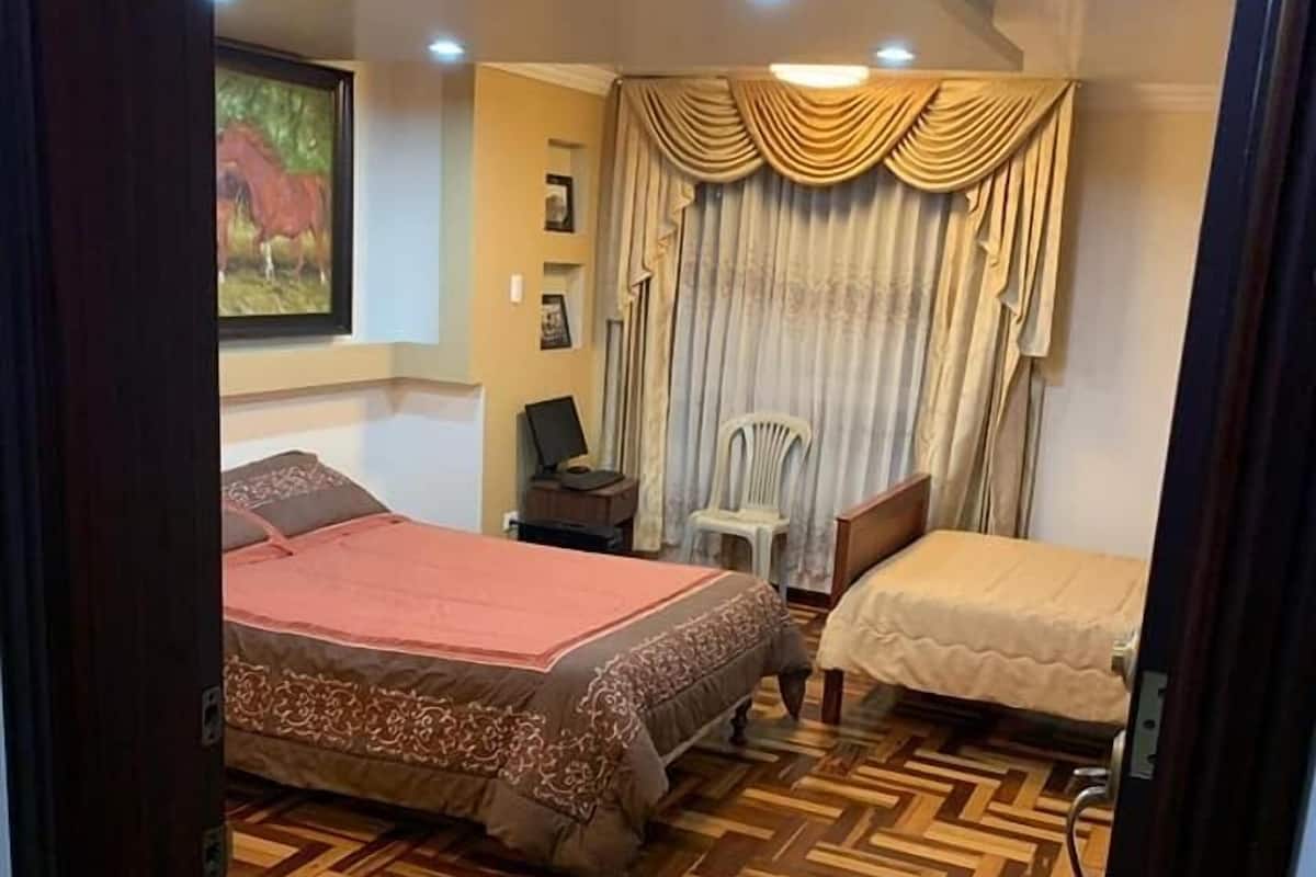 Un dormitorio con una cama grande, una cama más pequeña, un escritorio con silla, una lámpara y un cuadro en la pared.