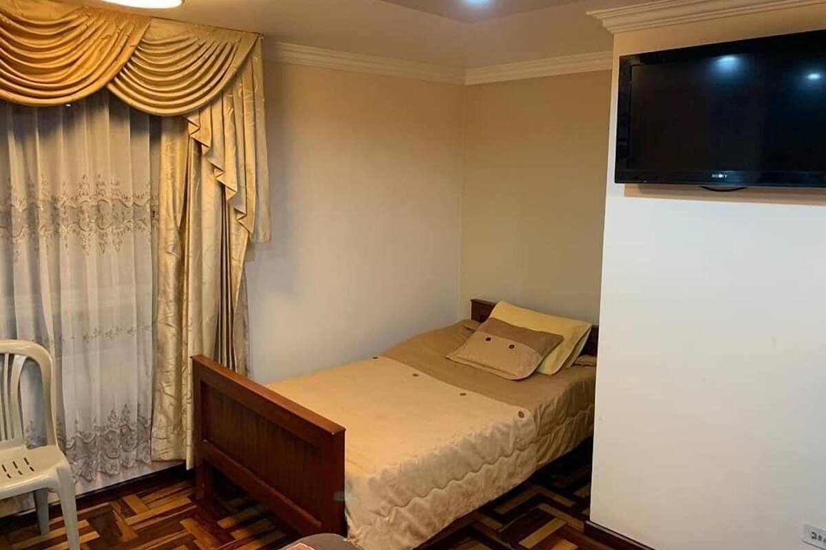 Una habitación pequeña con una cama individual, un marco de cama de madera, una silla blanca, un televisor montado en la pared y una cortina estampada.