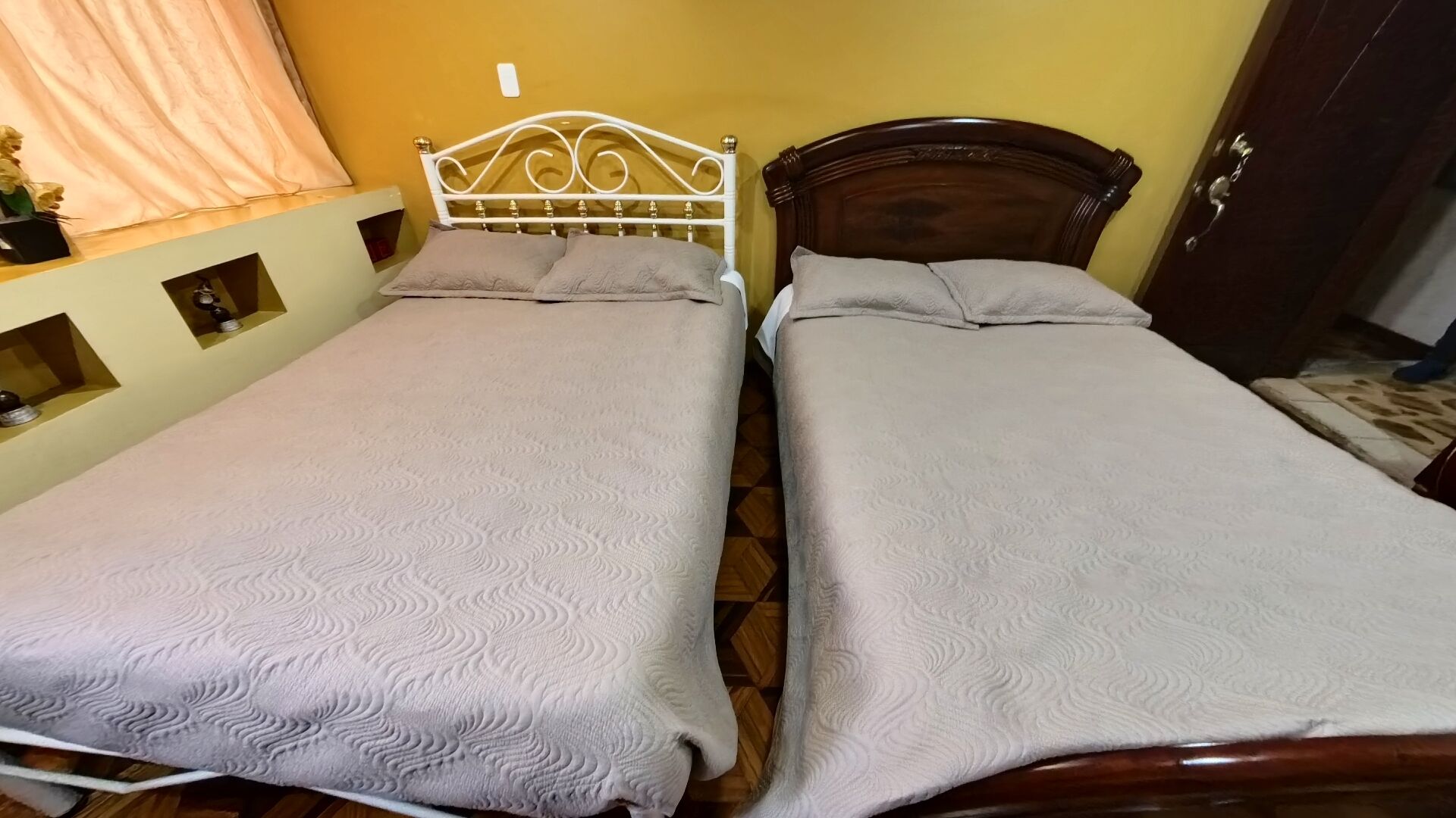Two single beds with wooden frames, a small shelf with decorative items, and a door to the right.