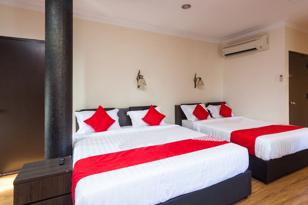 Habitación de hotel con dos camas, cada una con ropa blanca y almohadas rojas, una mesita de noche de madera, un pilar grande y un equipo de aire acondicionado en la pared.