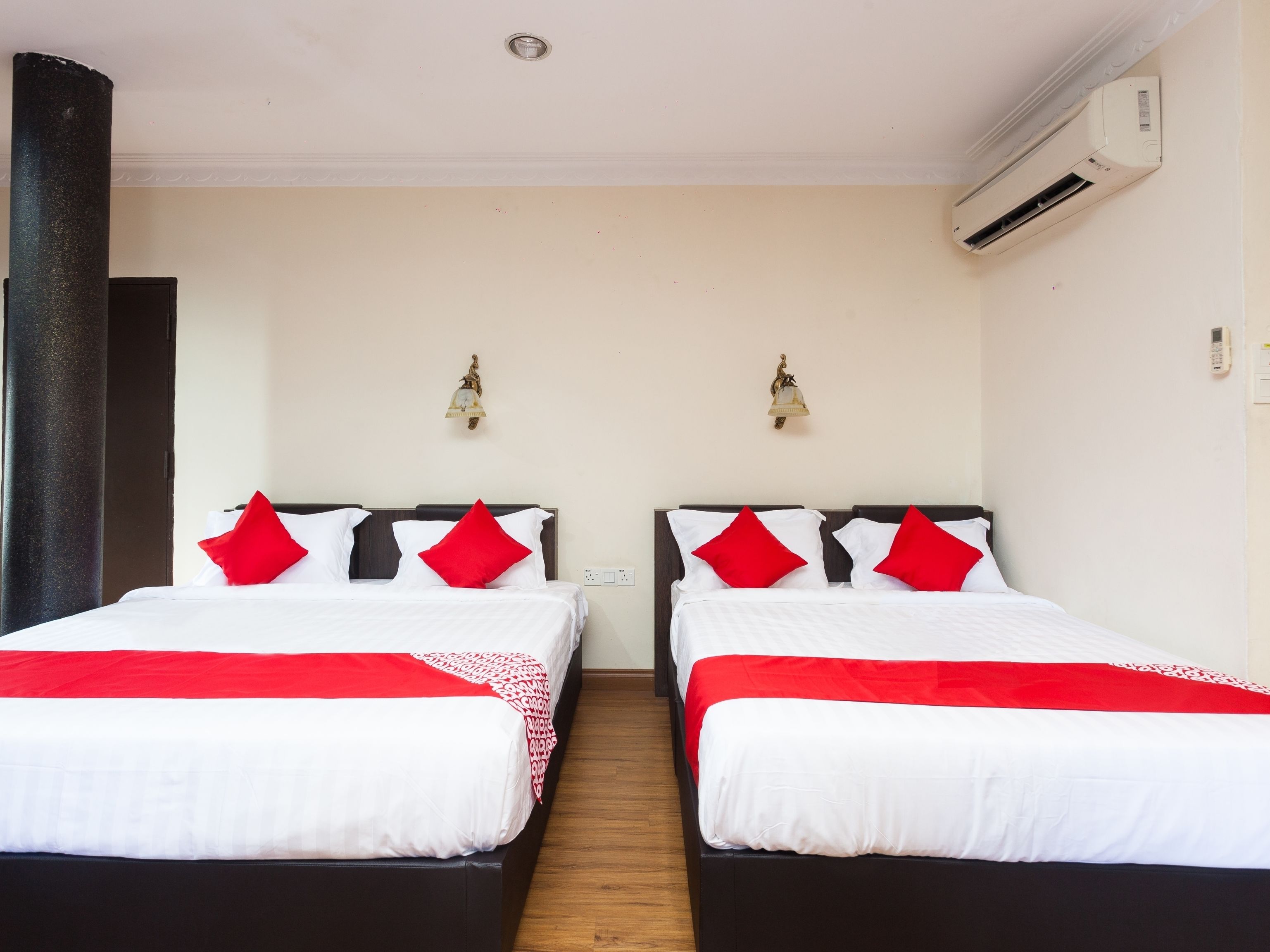 A hotel room with two beds, each with white bedding and red pillows, a wooden floor, and an air conditioning unit on the wall.