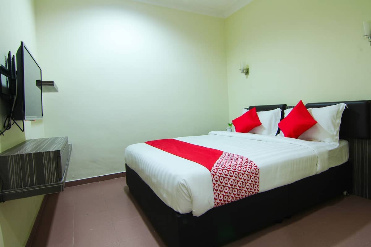 Una cama bien tendida con una colcha blanca y roja, un cabecero negro y dos almohadas rojas. A la izquierda de la habitación hay una mesita con un televisor.