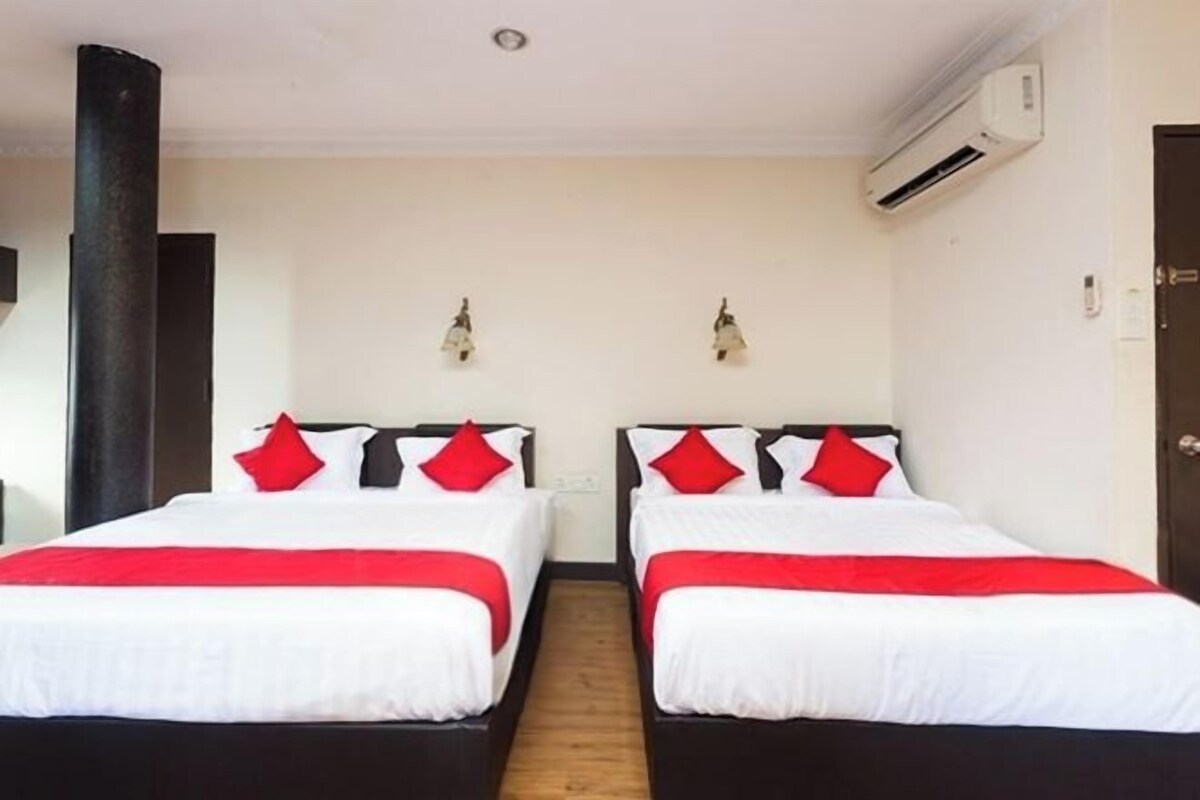 Quarto de hotel com duas camas, cada uma com roupa de cama branca e almofadas vermelhas, chão de madeira, ar condicionado na parede e uma porta à direita.
