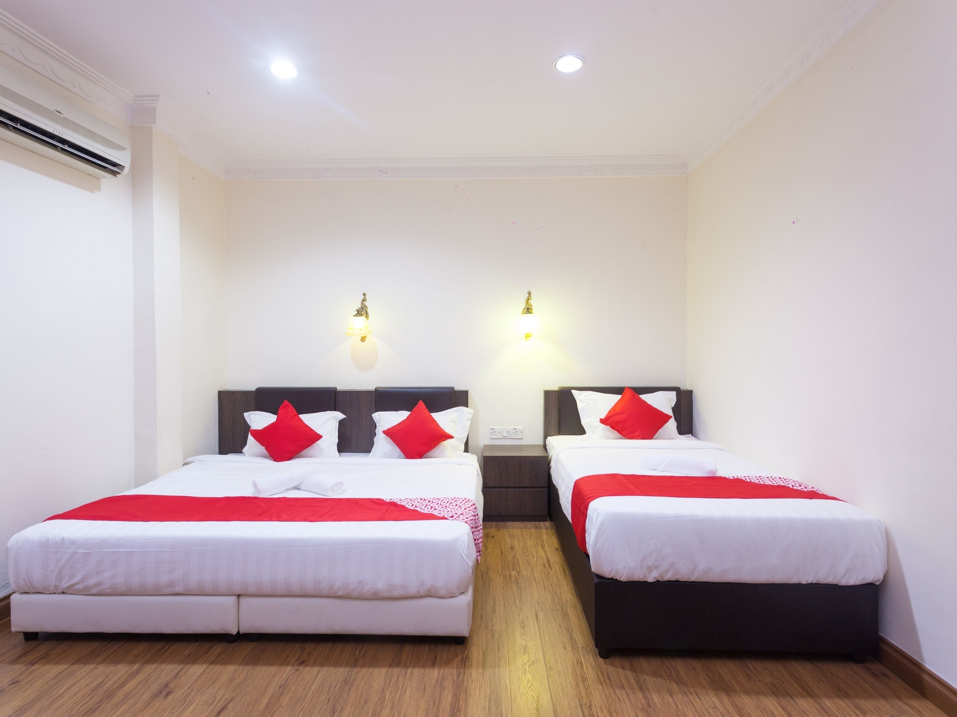 A hotel room with two single beds, each with a wooden headboard, white bedding, and red pillows. There are two wall-mounted light fixtures and a ceiling light.