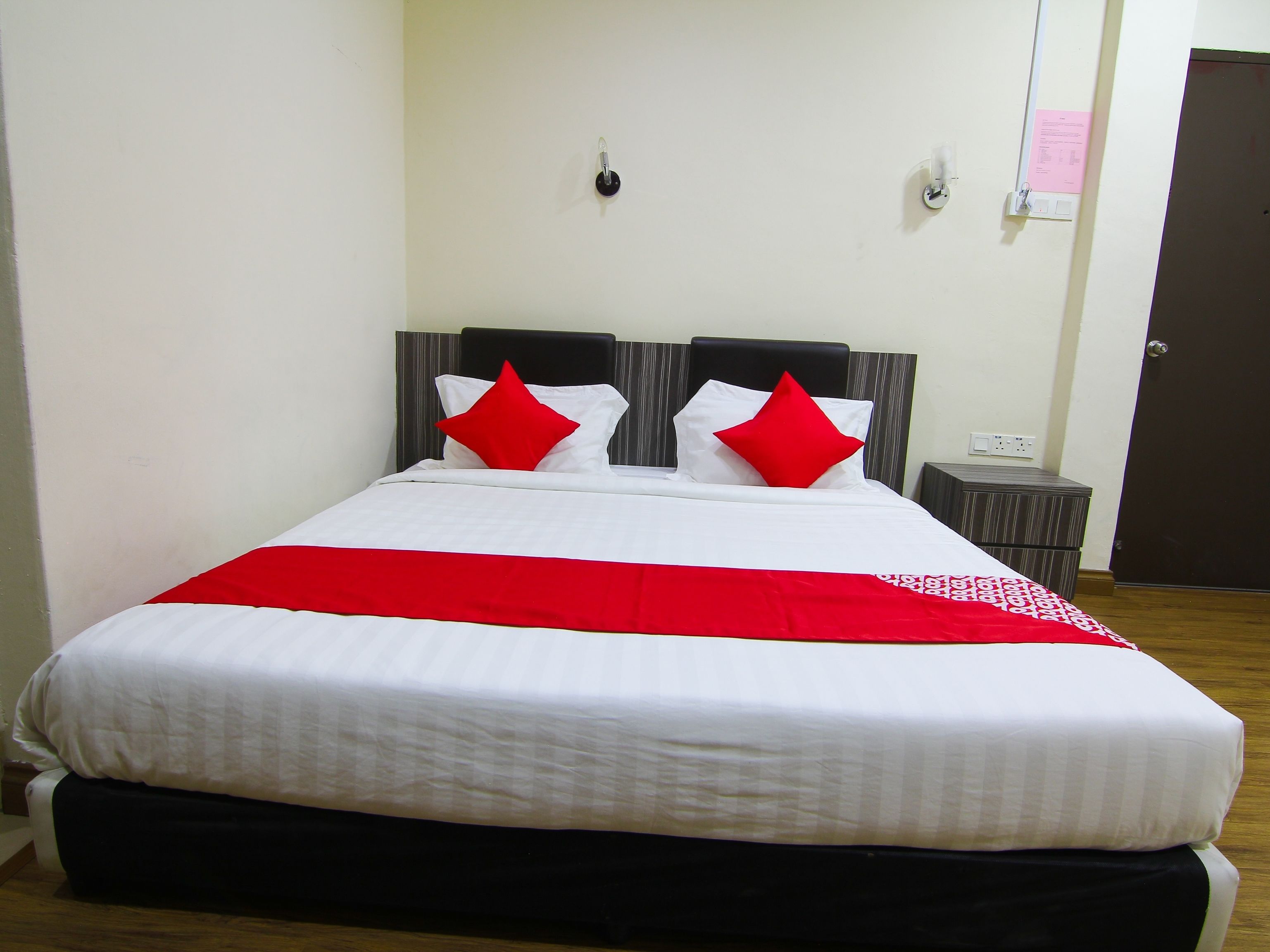 A neatly made bed with white bedding and red accents, a small bedside table, and a door in the background.
