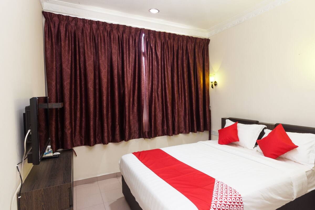 Una habitación de hotel con una cama grande, ropa de cama roja y blanca, un televisor sobre un soporte de madera y una ventana con cortinas oscuras.