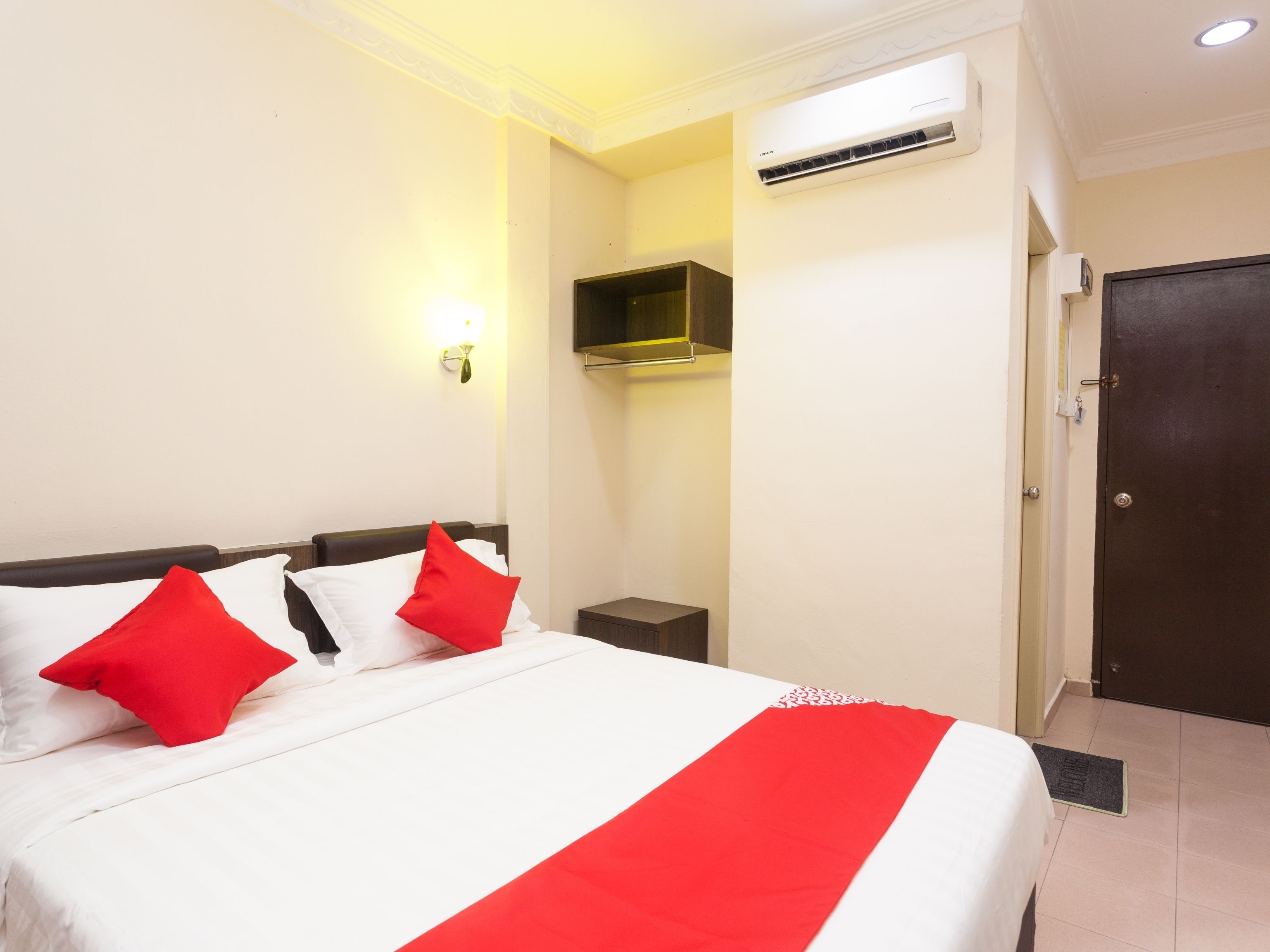 A hotel room with a bed, a nightstand, an air conditioning unit, a wall-mounted shelf, and a door.