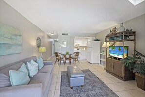 A living room with a sofa, ottoman, and a television on a cabinet. There is a dining area with a table and chairs, and a kitchen in the background. - Idyllic Inland Condo | Pool & Beach Access (Marco Island)