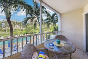 Een balkon met rieten meubels, een glazen tafel en uitzicht op een zwembad en palmbomen.