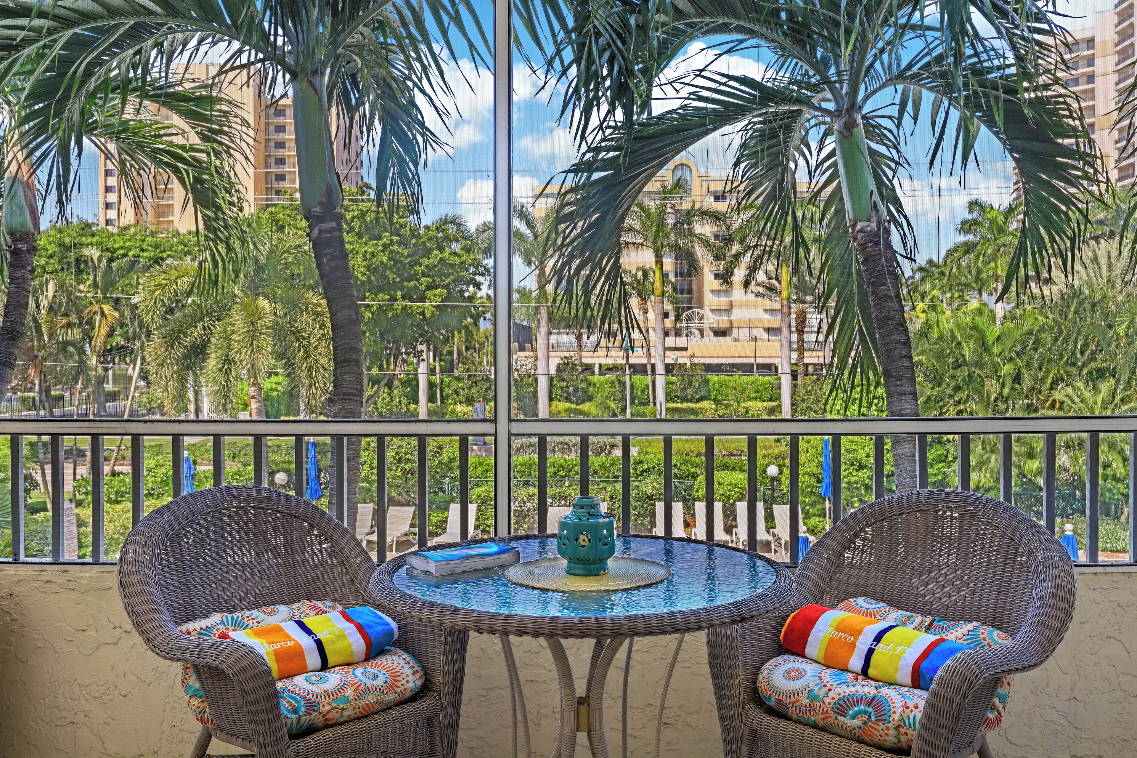 A balcony with wicker chairs and a table, overlooking a cityscape with palm trees and buildings.