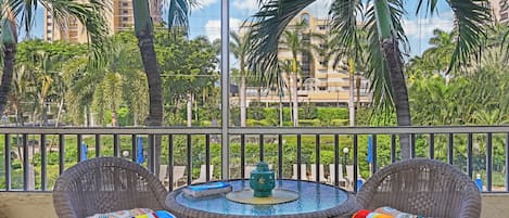 A balcony with wicker chairs and a table, overlooking a cityscape with palm trees and buildings.