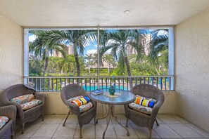 A balcony with wicker furniture, a small table, and a view of a pool and palm trees. - Idyllic Inland Condo | Pool & Beach Access (Marco Island)