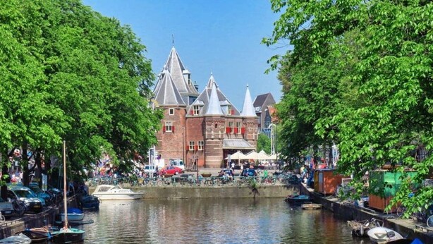 Un edificio histórico con múltiples torres, un canal con barcos y exuberantes árboles verdes enmarcan la escena.