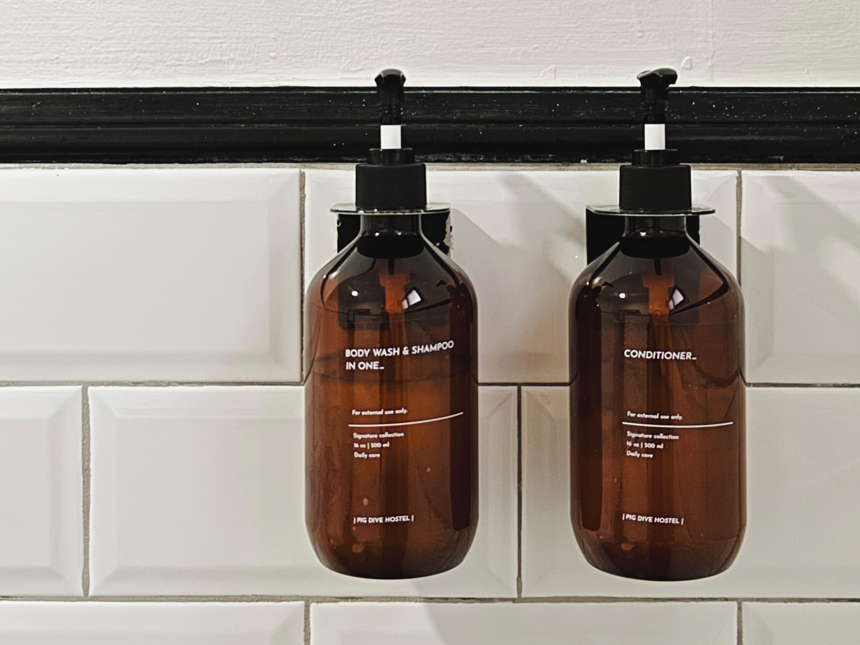 Two brown bottles with black pumps, labeled "BODY WASH & SHAMPOO" and "CONDITIONER," hanging on a white tiled wall.