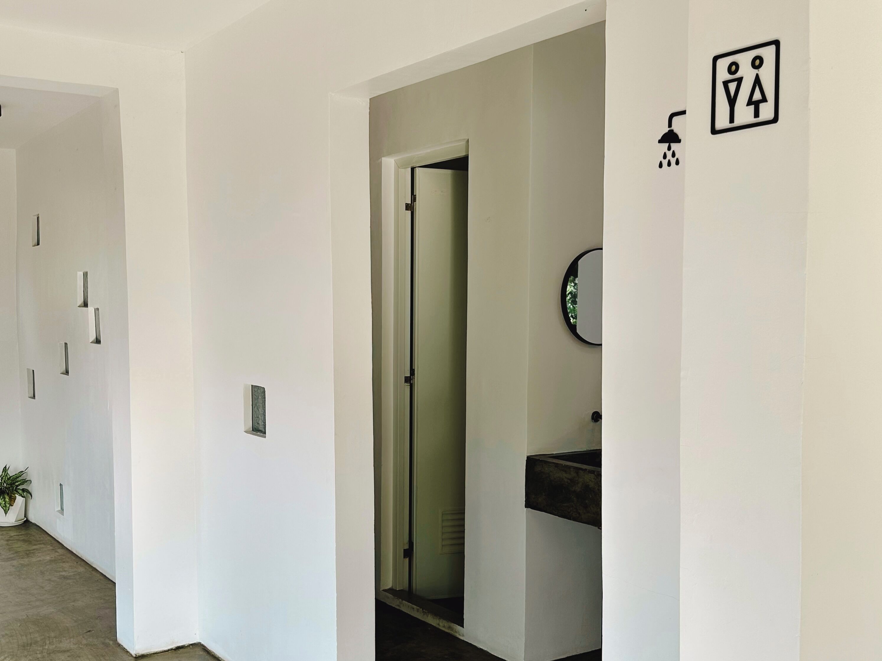 Un couloir avec un mur blanc, une plante en pot et une porte donnant sur une salle de bain équipée d’un lavabo et d’un miroir.