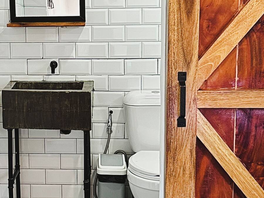 Un baño con pared de azulejos blancos, lavamanos de madera, inodoro blanco y puerta de madera.