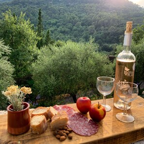 Una mesa con alimentos y bebidas, incluyendo pan, queso, manzanas, almendras y una botella de vino, con un paisaje de colinas boscosas de fondo.