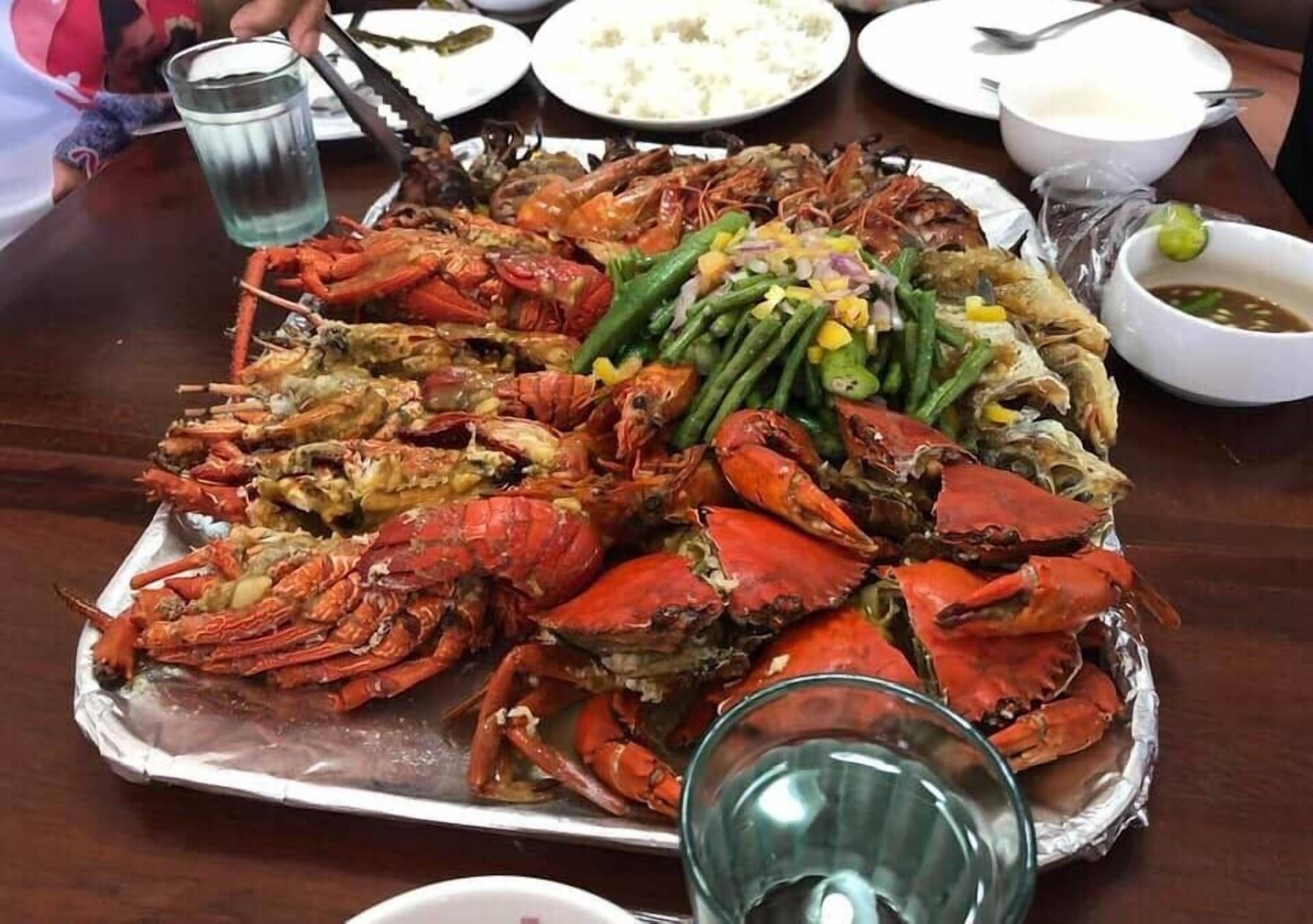 Una bandeja grande de mariscos cocidos, incluyendo cangrejos y pescado, acompañada de judías verdes y un recipiente pequeño con salsa.