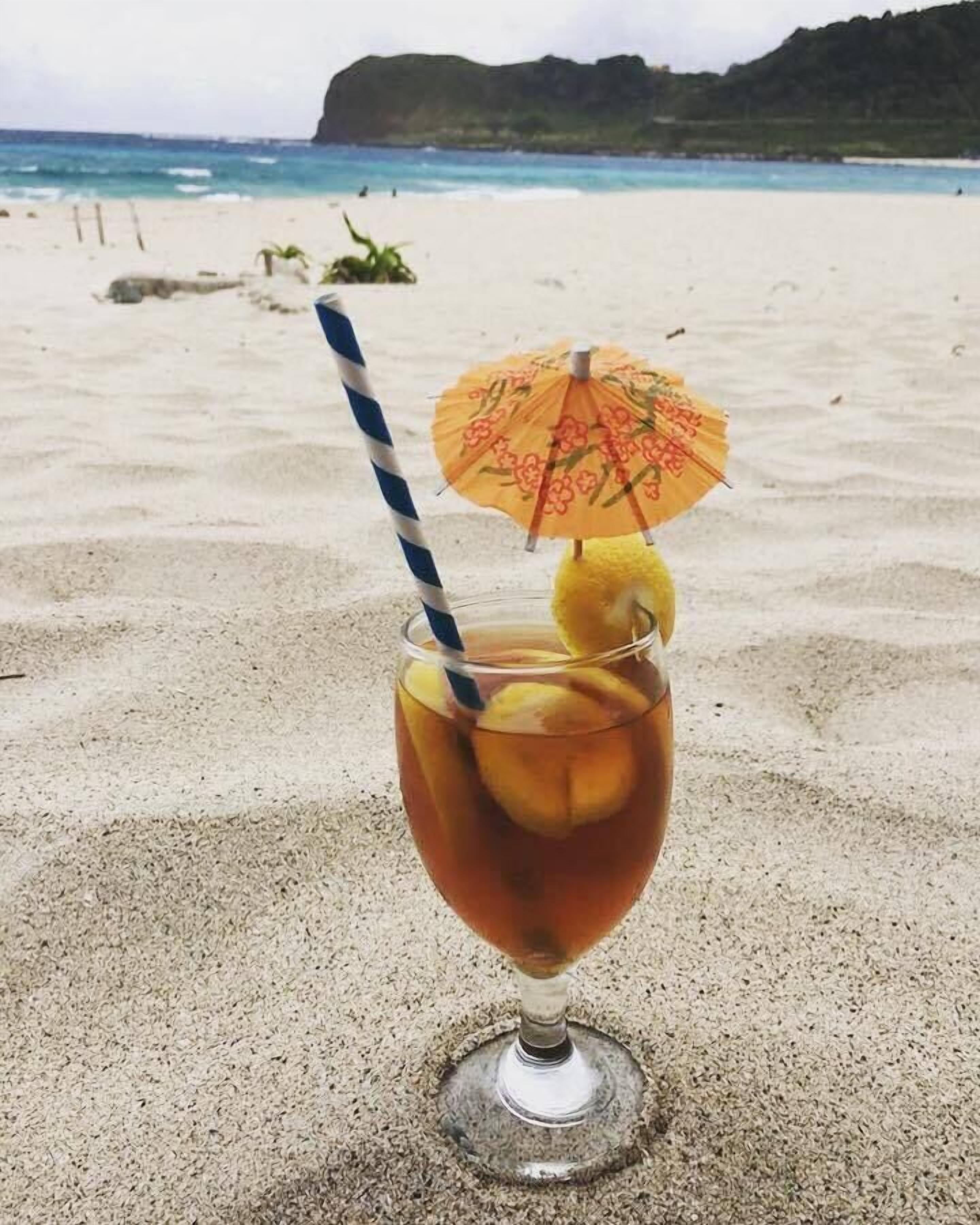 Un vaso de cóctel con pajita y sombrilla en una playa arenosa, con vista al océano y colinas al fondo.