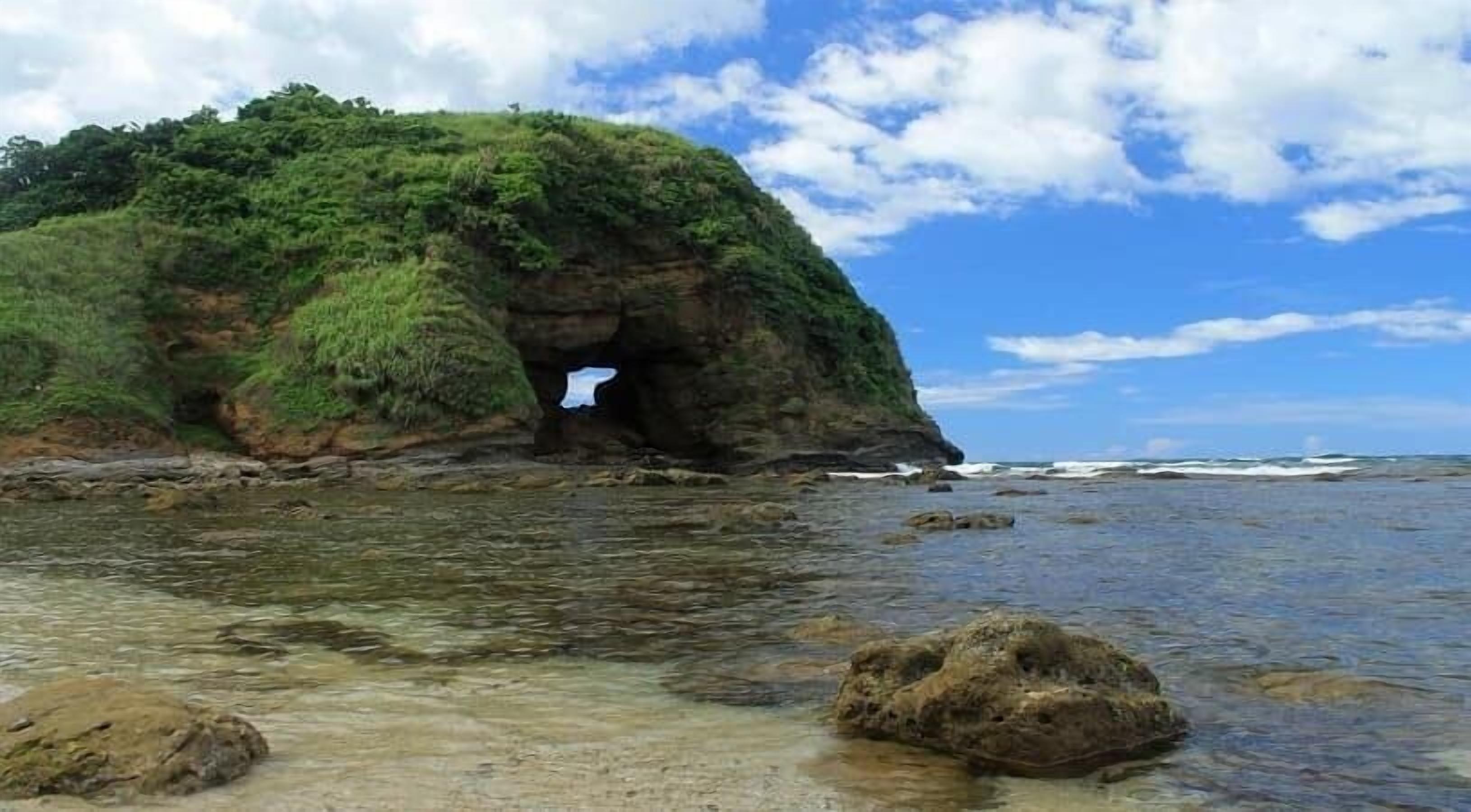 Un paisaje costero con una formación rocosa destacada que presenta la entrada de una cueva, aguas claras y poco profundas, y un cielo parcialmente nublado.