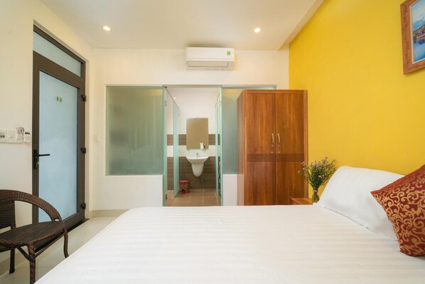 A hotel room with a bed, a wooden wardrobe, a bathroom visible through a glass door, and a small table with a vase of flowers.
