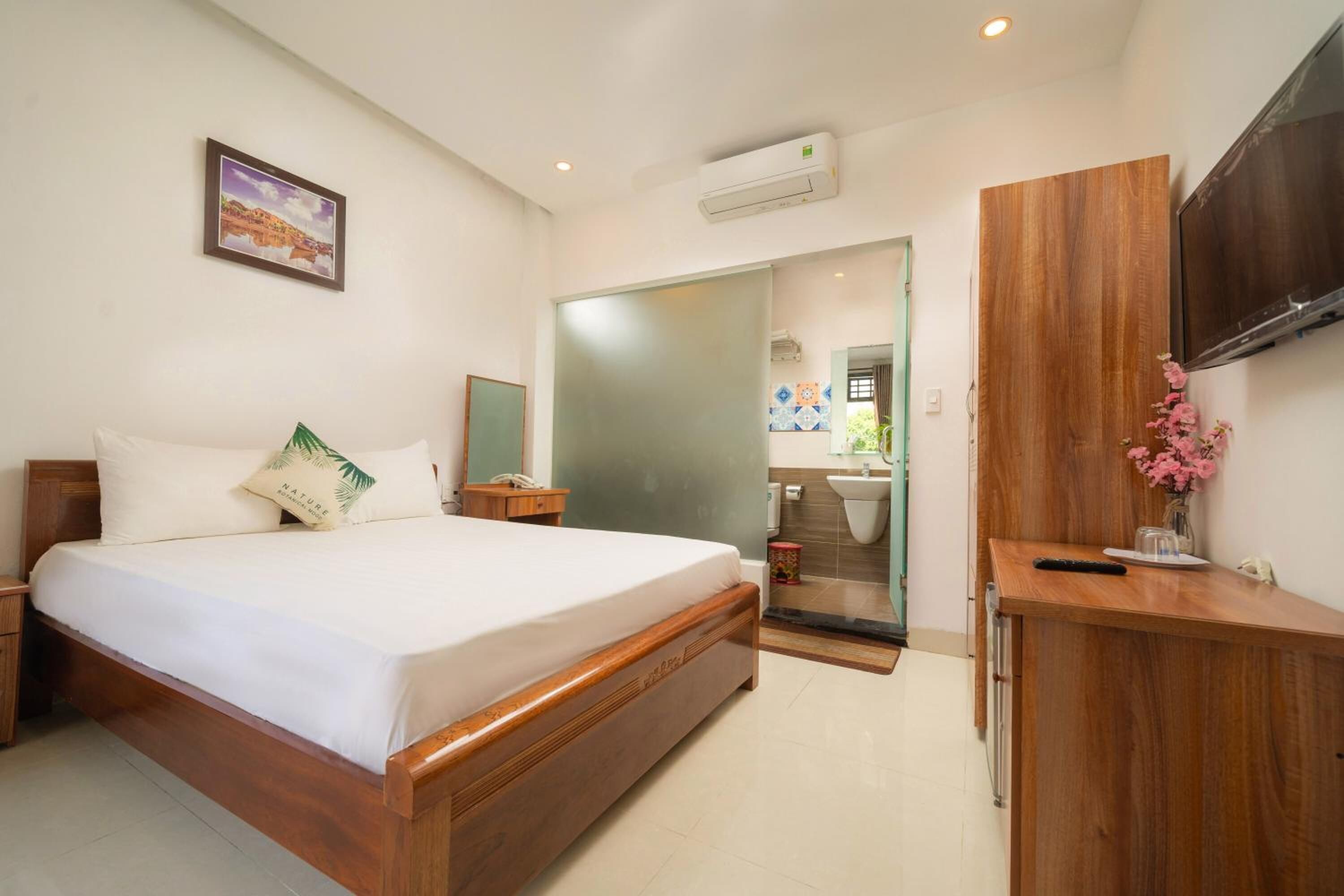 Un dormitorio con cama de madera, un escritorio, un televisor y un baño visible a través de una puerta de cristal.