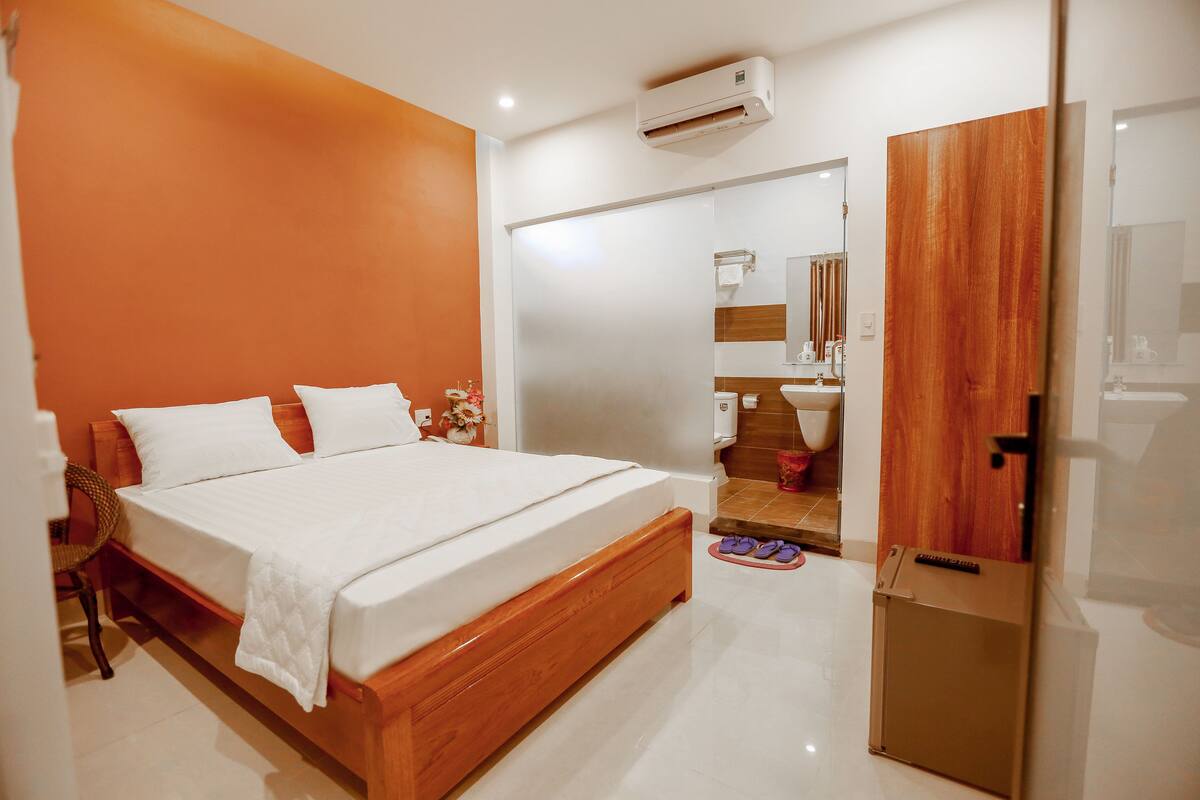 A modern bedroom with a wooden bed, white linens, a small wooden cabinet, and a bathroom visible through a glass door.